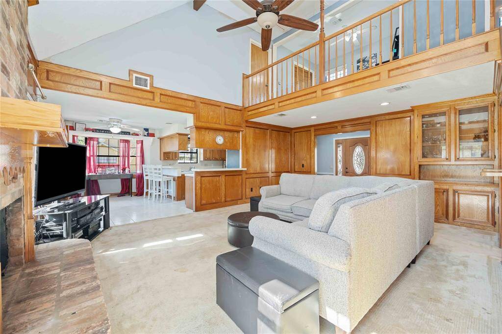 This inviting living space features a spacious open layout with a high ceiling and a cozy brick fireplace. The room extends to a kitchen with wooden cabinetry and a breakfast bar. A loft area overlooks the living room, adding charm and potential for additional space.