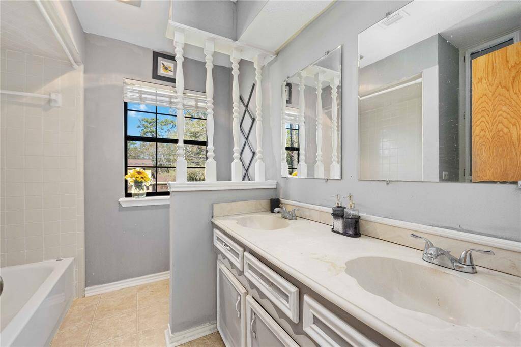This bathroom features a dual-sink vanity with ample storage, light gray walls, and a large mirror. It has a window with a view, adding natural light, and a tiled bathtub area. The space feels bright and functional.