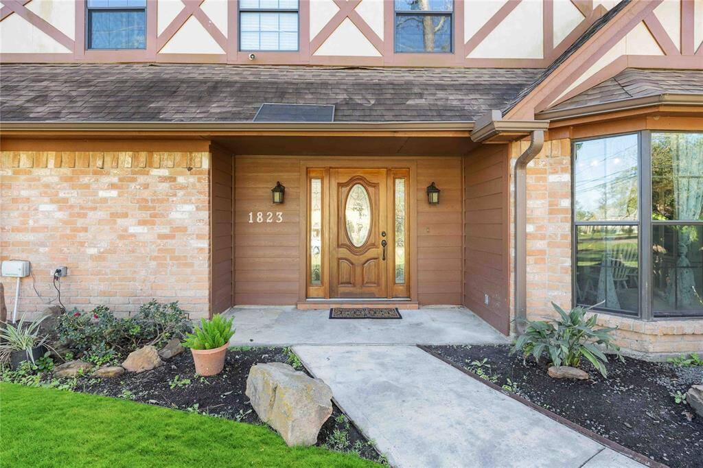 This photo showcases a welcoming entrance to a home with a wooden front door, flanked by brick and siding. It features a small garden area with plants and a paved walkway leading to the door. The house number "1823" is visible, and there are large windows on the right side.