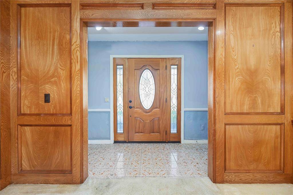 This photo showcases a welcoming entryway with rich wooden paneling and a decorative front door featuring glass accents. The tiled floor adds elegance, and the soft blue walls offer a calming touch.