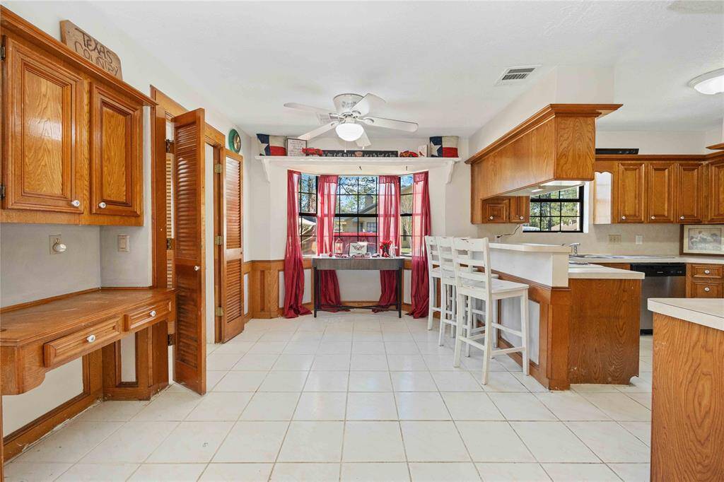This kitchen features warm wooden cabinetry, a breakfast bar with high chairs, and a cozy dining nook by the window with red curtains. The space is bright, thanks to ample natural light, and has a tiled floor for easy maintenance.