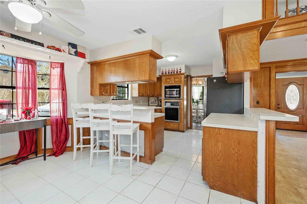 This spacious kitchen features warm wood cabinetry, white countertops, and a breakfast bar with seating. A large window provides natural light, highlighting the open layout and modern appliances, including a built-in oven. The adjacent entryway and dining area add to the home's inviting atmosphere.