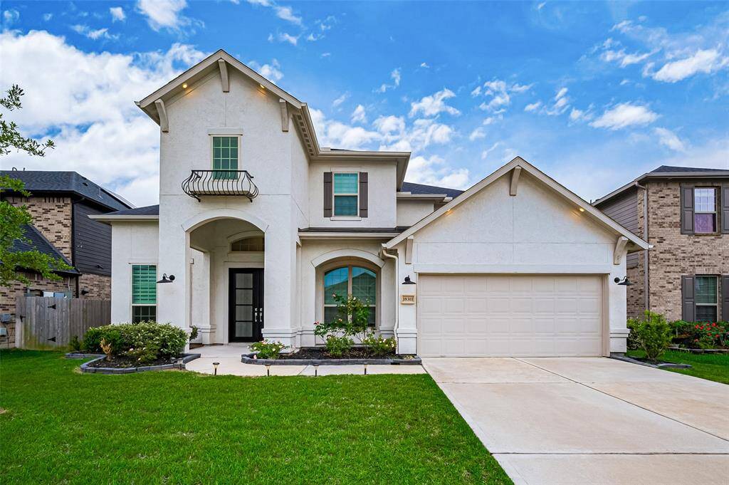 An elegant stucco façade is artfully accented by recessed lighting beneath the rooflines, creating a warm, inviting glow after dusk. The grand arched entryway showcases stately double doors, framed by manicured plant beds. A wide, smooth driveway leads to the two-car attached garage, flanked by a generous green lawn with a young, flourishing tree.