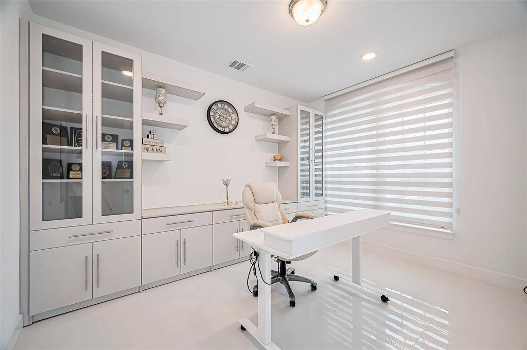 A quiet, inspiring workspace awaits with a large window and an expansive built-in wall unit that includes lower cabinets, open shelving, and upper glass-front storage—offering both form and function.
