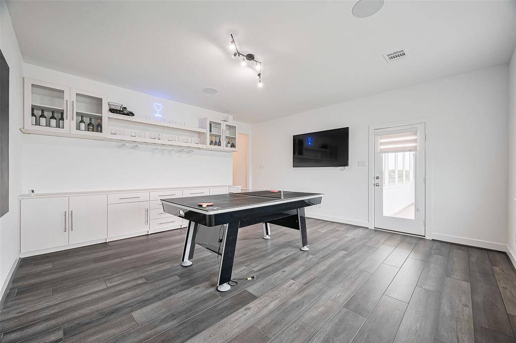 A large and inviting upstairs game room with smooth dark flooring and a striking light fixture. A mounted overhead bar cabinet offers a polished touch for casual entertaining.
