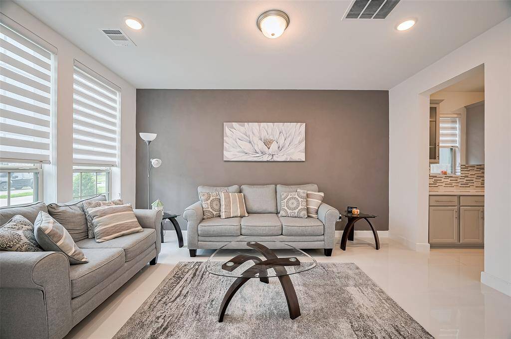 Bathed in natural light from oversized windows, this serene sitting space features a soft gray accent wall and flows effortlessly into the kitchen through an open doorway—ideal for relaxed entertaining.