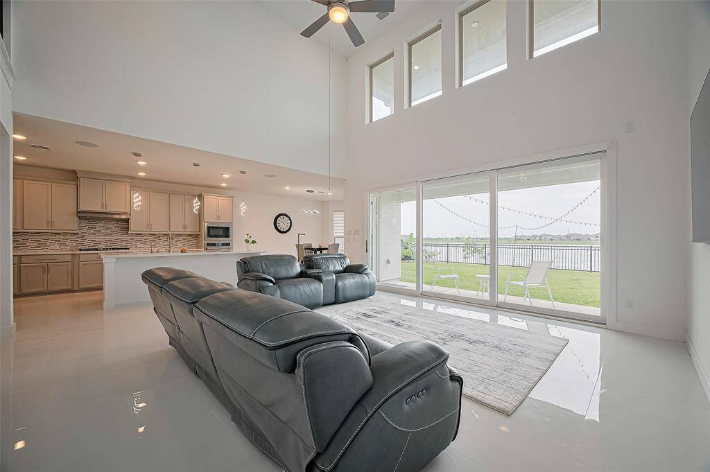 A stunning open-concept living space unfolds with dramatic two-story ceilings and a full wall of windows, flooding the room with sunlight. A sleek ceiling fan adds comfort, while polished white marble flooring reflects the light for an airy, upscale ambiance.