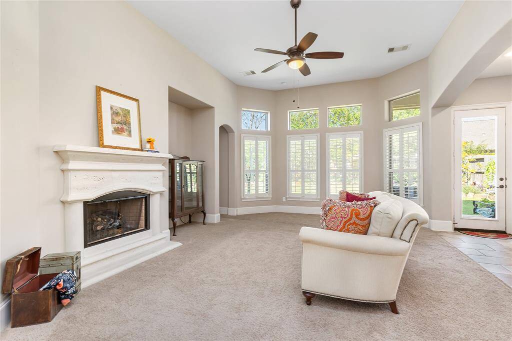 This spacious living room features a cozy gas fireplace, large windows with shutters letting in ample natural light, and a ceiling fan. The neutral carpet and walls create a warm, inviting atmosphere. The room opens to outdoor views through a glass door, adding to its charm.