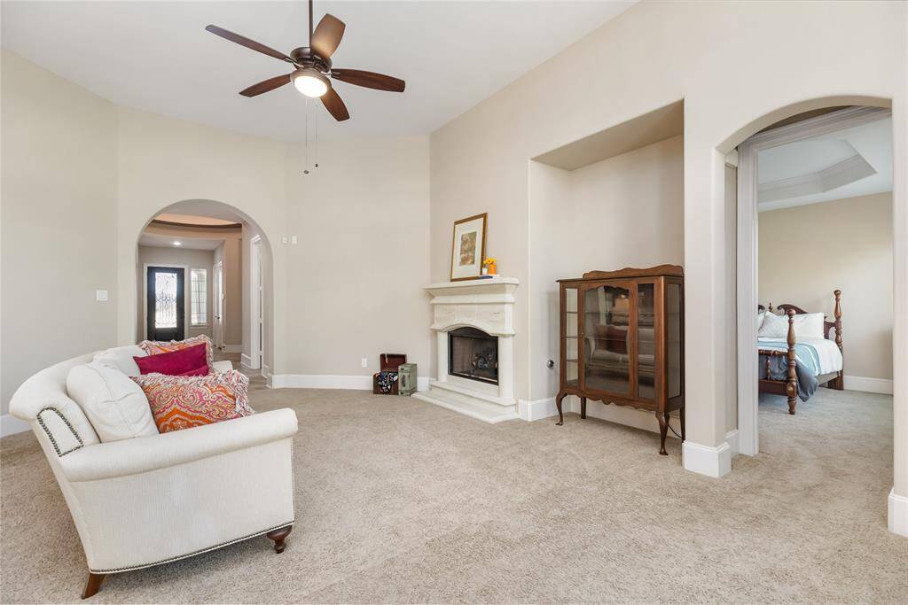 This spacious living area features a cozy fireplace, soft carpet flooring, and neutral walls. It offers elegant archways leading to adjacent rooms and a ceiling fan, creating a welcoming atmosphere.