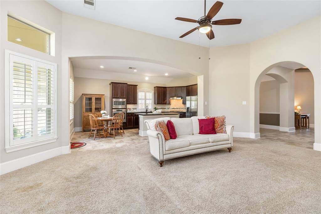 This spacious open-concept living area features a cozy carpeted section with a ceiling fan, leading into a bright kitchen with dark wood cabinets and granite countertops. Large windows with shutters let in natural light, and there's a convenient dining nook adjacent to the kitchen. Arched doorways add architectural interest, enhancing the home's inviting atmosphere.