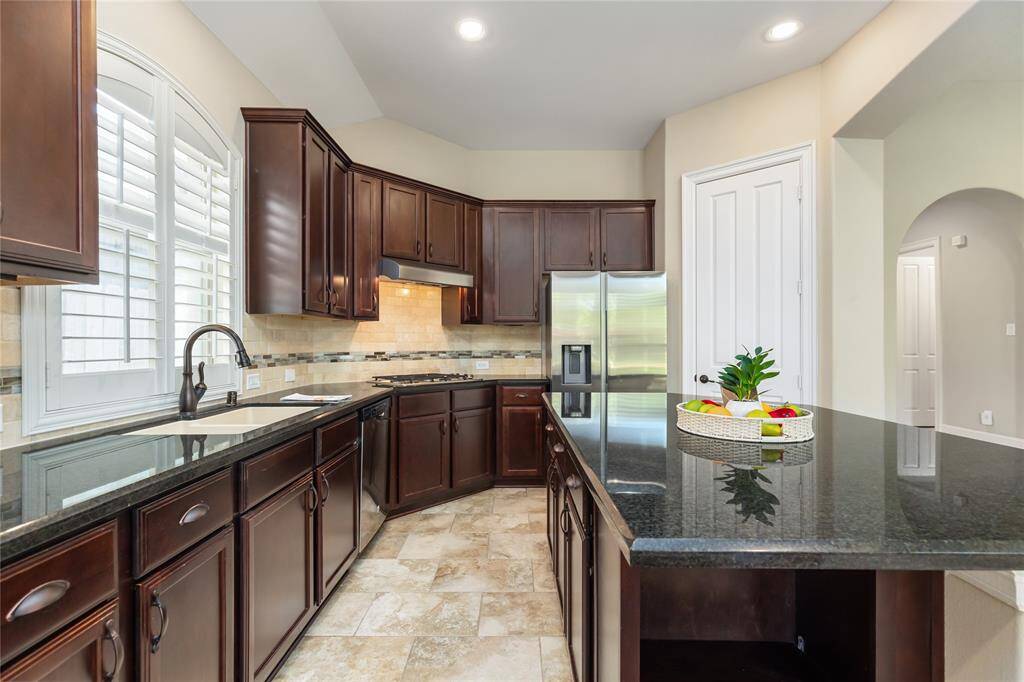 This kitchen features elegant dark wood cabinets, a spacious granite countertop, stainless steel appliances, and a large island. The neutral tile flooring and ample natural light from the arched window create a warm and inviting space.