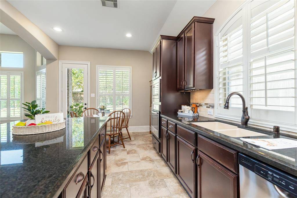Bright kitchen with modern dark wood cabinets, granite countertops, and tile flooring. Features a large island, stainless steel appliances, and ample natural light from windows and a glass door leading outside. Cozy dining area with a view of the landscaping.