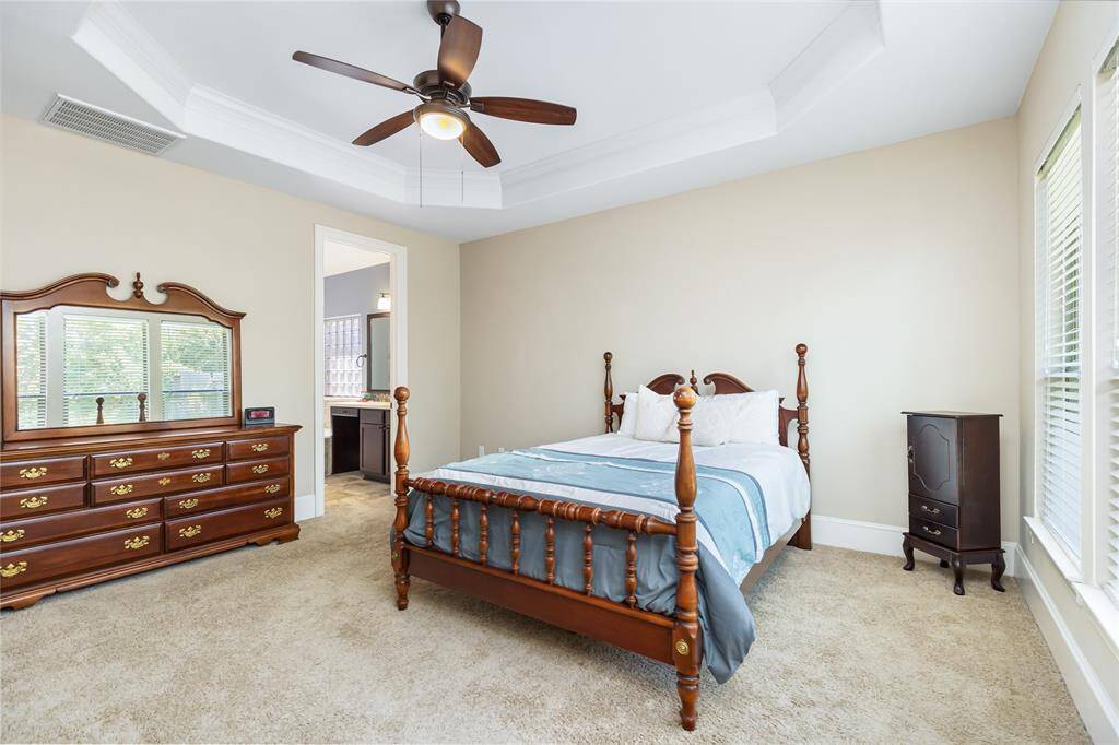 This is a spacious bedroom featuring a tray ceiling and a ceiling fan. It has a large window allowing natural light, and an adjoining bathroom. The carpeted floor adds a cozy touch.