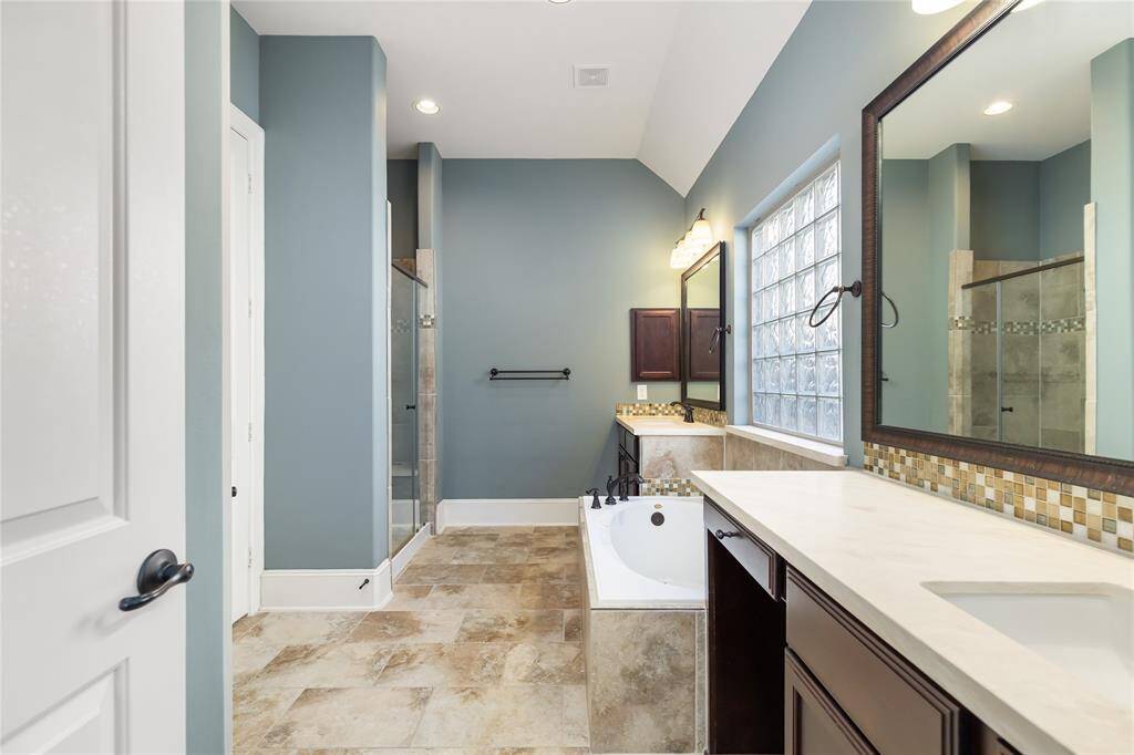 This primary bathroom features a modern design with a spacious layout. It includes a large glass-enclosed shower, a deep soaking tub, elegant vanity with dual sinks, oversized closet and stylish tile flooring. Natural light fills the space through a frosted window, creating a serene ambiance.