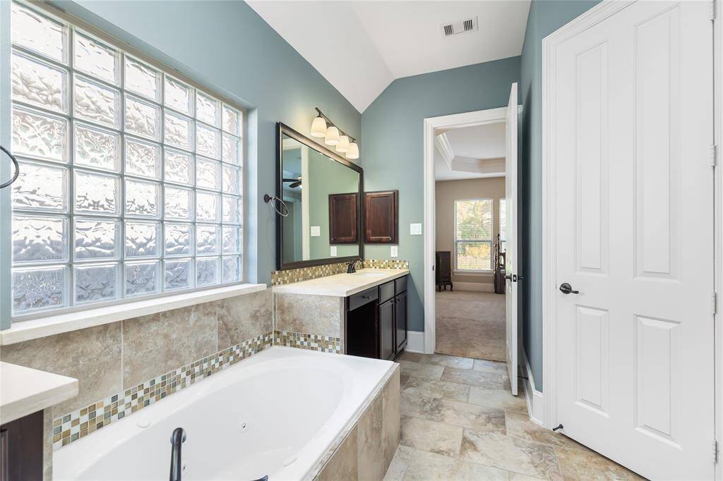This primary bathroom features a spacious layout with a large soaking tub, stylish tile accents, and a glass block window for natural light. It includes a dual sink vanity with dark wood cabinetry and modern lighting. The room leads into a primary bedroom, adding convenience and comfort.