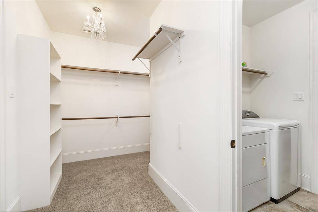 This photo shows a spacious walk-in closet with built-in shelves and hanging rods, featuring carpeted flooring and a small chandelier. Adjacent is a laundry area, adding convenience to the space.