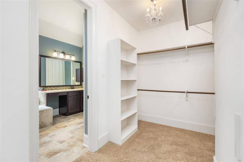 This photo shows a spacious walk-in closet with built-in shelving, hanging rods, and a carpeted floor, leading into a bathroom with a large mirror, modern lighting, a dark vanity, and tiled flooring.