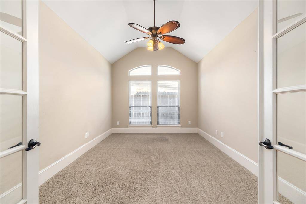 Bright and spacious room with vaulted ceilings, neutral walls, and plush carpet. Features a ceiling fan and large window for natural light, accessible through elegant French doors. Can also be used as a game room, bedroom or office.