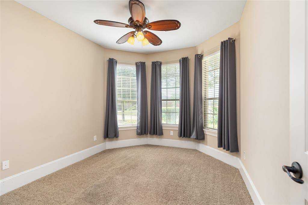 This bedroom features a cozy, carpeted space with neutral walls and a ceiling fan with wooden blades. It has large, bay windows adorned with dark curtains, offering ample natural light.