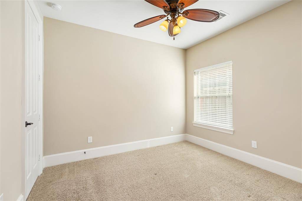 This bedroom features neutral walls, a cozy carpeted floor, and a ceiling fan with lights. There's a window with blinds for natural light and a white door, offering a simple and versatile space.