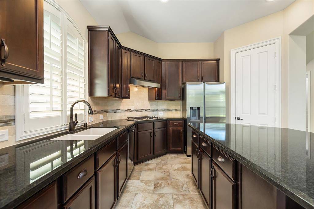 This kitchen features elegant dark wood cabinetry, granite countertops, and stainless steel appliances. It has a spacious layout with ample storage, a modern tile backsplash, and natural light from large windows with plantation shutters.