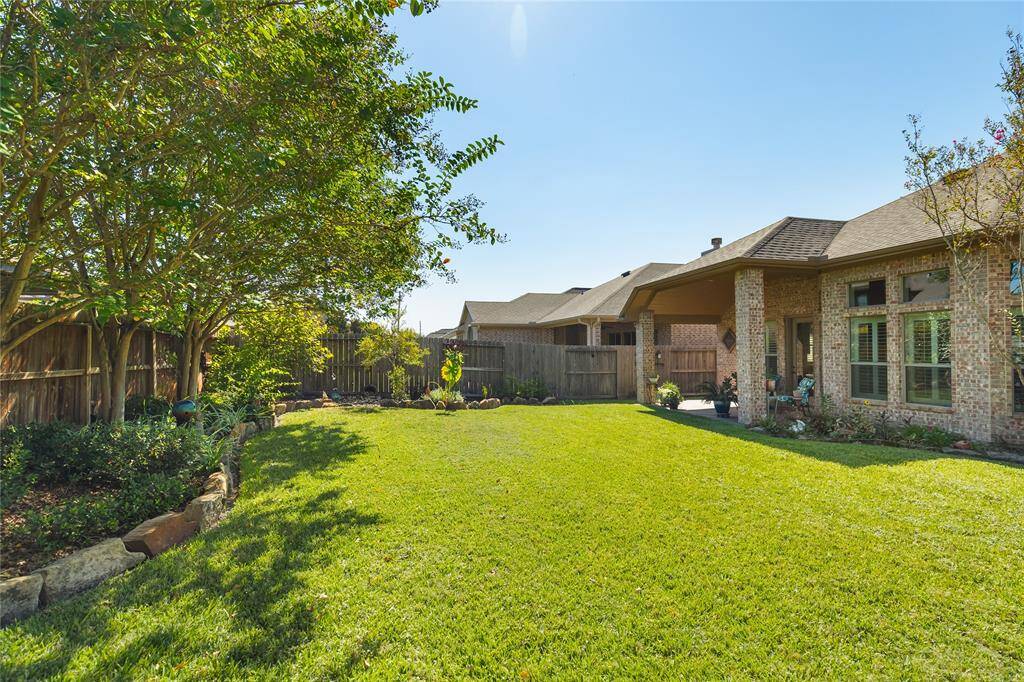 This photo showcases a spacious backyard with a well-maintained lawn, bordered by mature trees and a wooden fence. The brick exterior of the house features large windows and a covered patio area, providing a perfect spot for outdoor relaxation and entertainment.