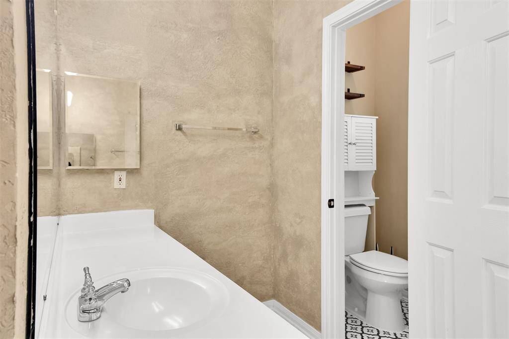 Features a bathroom with a textured beige wall, a white countertop sink, a mirror, and a separate toilet area with built-in shelving. The space is compact and functional.