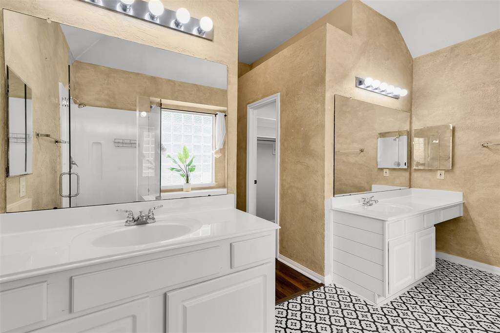 This bathroom features dual vanities with sleek white countertops, large mirrors, and modern lighting. The space is highlighted by textured beige walls and patterned tile flooring. A glass-enclosed shower and a window with frosted glass provide both functionality and natural light.