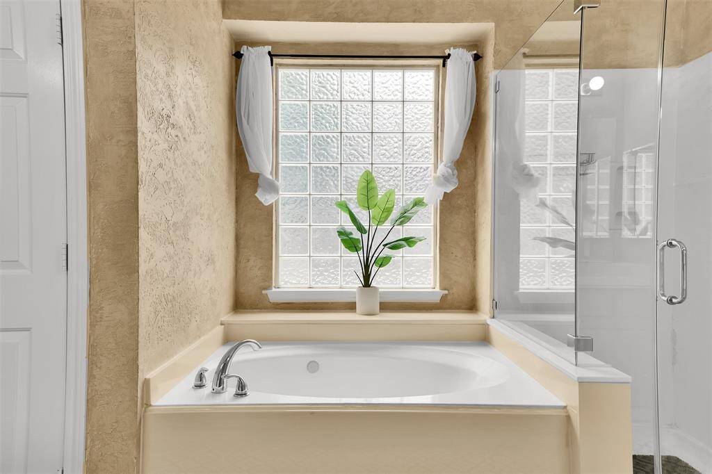This bathroom features a luxurious soaking tub with sleek fixtures, set against textured walls. A large glass-block window provides natural light, and a glass-enclosed shower adds modern elegance.