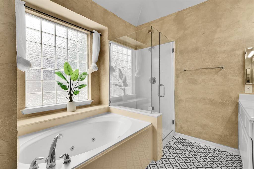 This bathroom features a spacious soaking tub next to a large glass-block window, providing natural light. It includes a sleek glass-enclosed shower and elegant patterned floor tiles, creating a modern and inviting space.