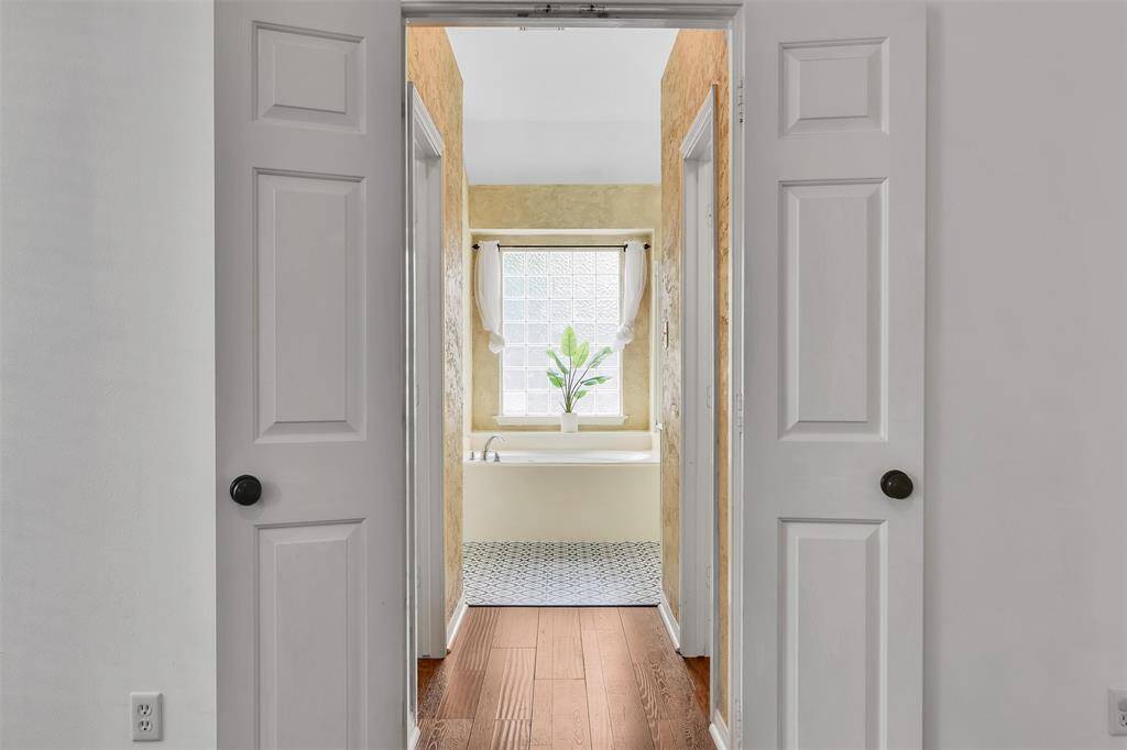This photo features a cozy bathroom entrance with white double doors. It opens to a bright space with a large window, textured walls, and a bathtub. The flooring transitions from wood to patterned tile.
