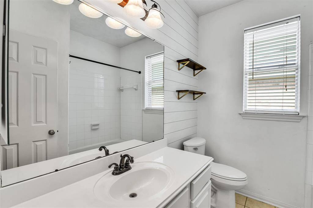 This bathroom features a clean, modern design with white cabinetry and walls, a large mirror, and stylish lighting. It includes a shower-tub combo and has natural light from a window with blinds. Two wooden shelves add a touch of warmth and functionality.