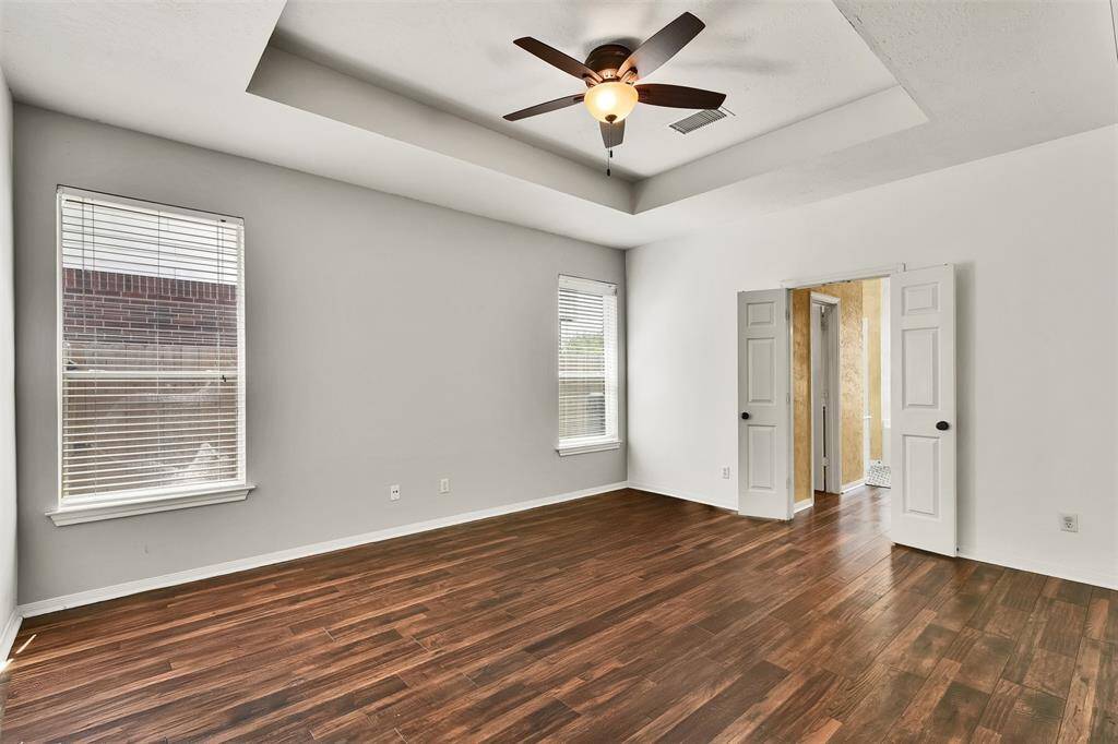 Bright, spacious room with wood flooring, two windows for natural light, a ceiling fan, and a tray ceiling design. Double doors open to an adjacent spa like on suite.