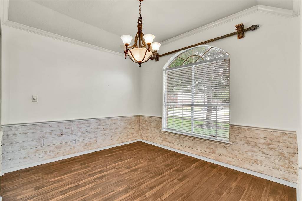 Features a large arched window with blinds, allowing plenty of natural light. It has wooden flooring and rustic wood paneling on the lower half of the walls. A decorative chandelier hangs from the ceiling, providing an elegant touch.