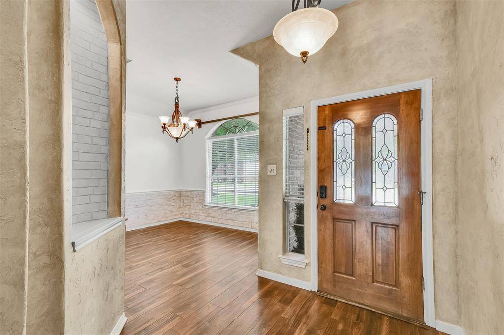 This inviting entryway features a wooden front door with decorative glass panels, warm-toned wood flooring, and textured walls. The adjacent dining area has a large window offering natural light and a stylish chandelier.