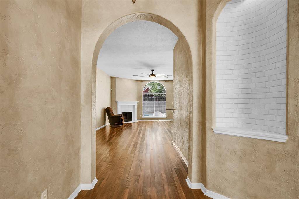 This showcases an inviting entryway with arched doorways, leading to a spacious living area. The room features wood flooring, a white brick accent wall, and a cozy fireplace. Natural light streams through large windows, enhancing the open, airy feel.
