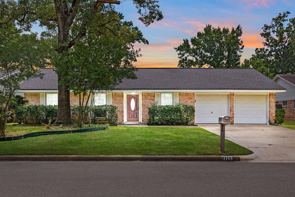 This charming single-story brick home features a well-maintained front yard with lush greenery and mature trees. It has a welcoming front door, a double garage, and a spacious driveway. The serene neighborhood setting is enhanced by a beautiful sunset backdrop.