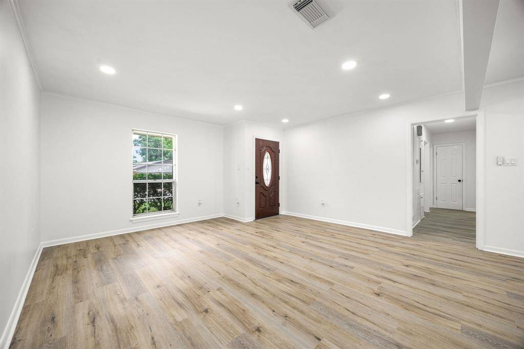 This spacious living room features modern recessed lighting, light wood flooring, and neutral walls, offering a bright and inviting atmosphere. A large window provides natural light, while the entryway includes a stylish wooden door.