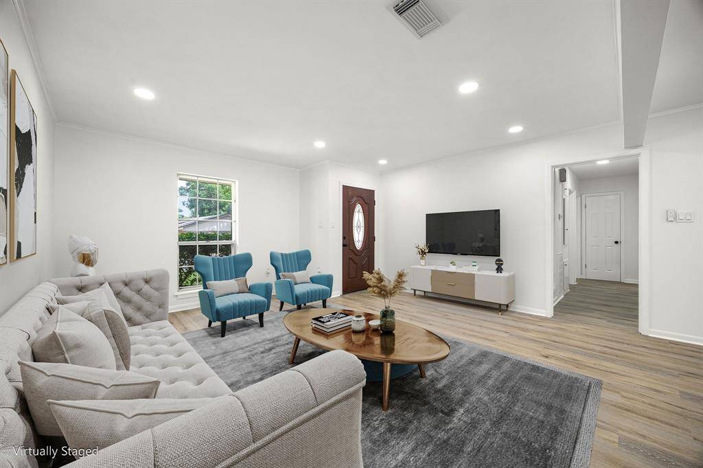 This photo showcases a bright, modern living room with light wood flooring, recessed lighting, and a large front window. The room is virtually staged with a comfortable sofa, two blue accent chairs, a coffee table, and a TV, creating an inviting space for relaxation and entertainment.