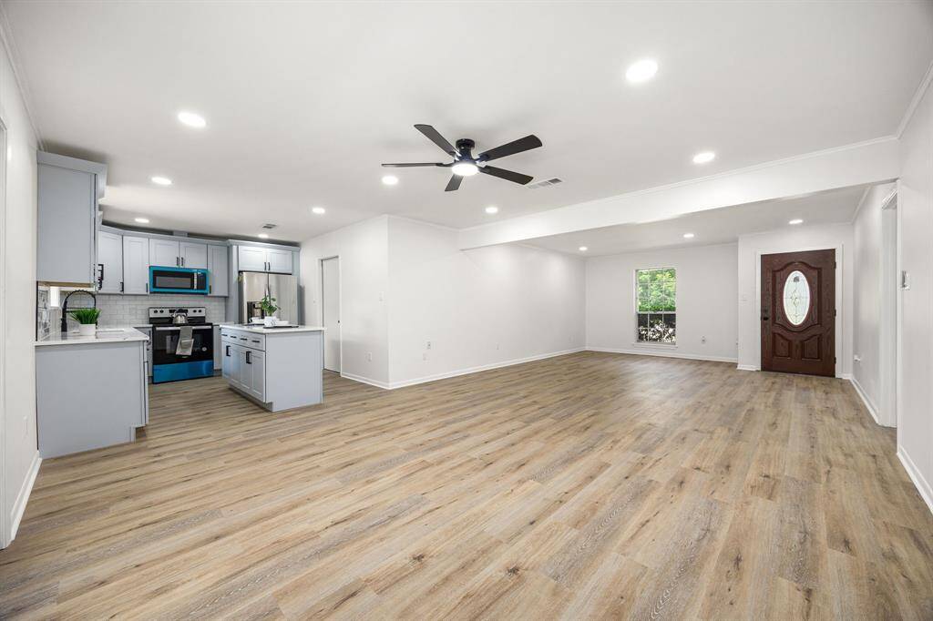 This photo showcases a spacious open-concept living area with light wood flooring, modern recessed lighting, and a stylish ceiling fan. The kitchen features sleek gray cabinets, stainless steel appliances, and a central island. Natural light fills the room through a large window, and the entrance has an elegant wooden door.