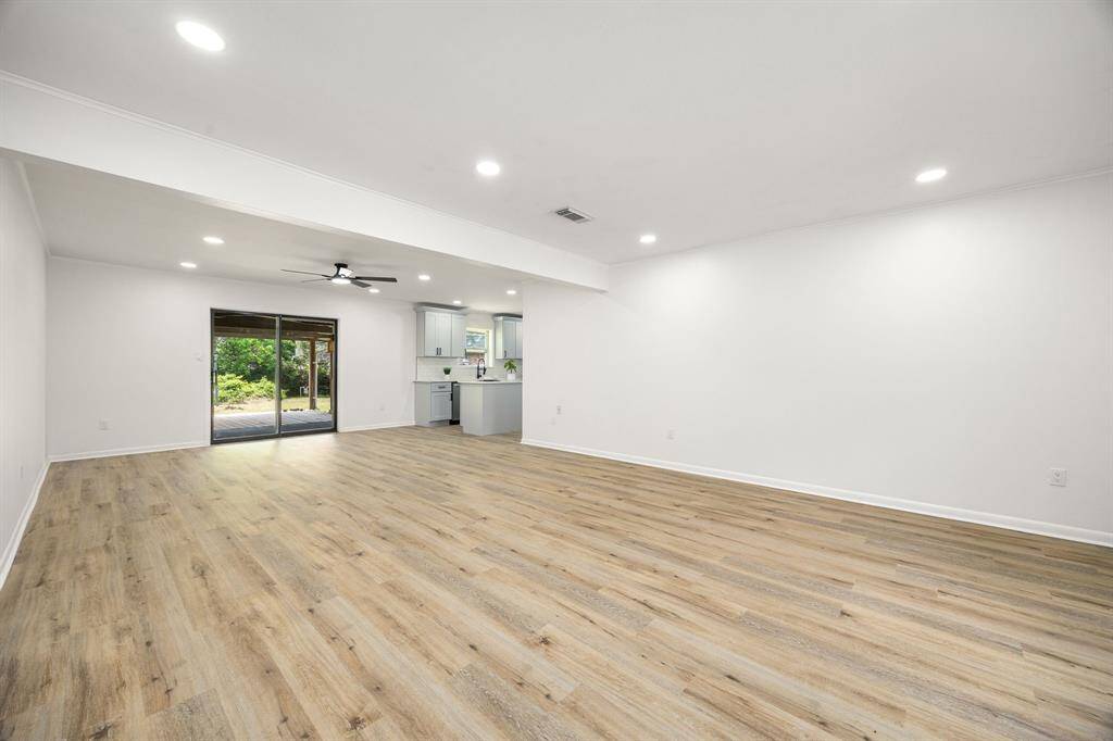 This spacious open-concept living area features sleek wood flooring, recessed lighting, and a ceiling fan. It seamlessly connects to a modern kitchen with white cabinetry and stainless appliances. Large sliding doors lead to a lush backyard, providing ample natural light.