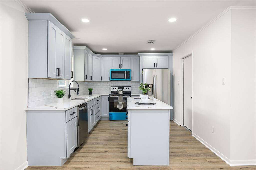 This modern kitchen features light gray cabinetry, stainless steel appliances, and a central island with seating. It has a bright, open layout with wood-style flooring and recessed lighting, creating a welcoming atmosphere.