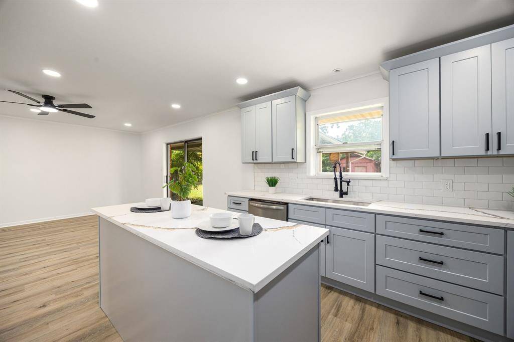 This kitchen features modern light gray cabinetry, a sleek island with a marble countertop, and a spacious, open layout. It has a large window for natural light, wood flooring, and recessed lighting, creating a bright, inviting space.