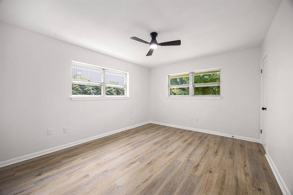 This room is bright and spacious with light-colored walls and wood flooring. It features two large windows allowing natural light and a modern ceiling fan for ventilation.