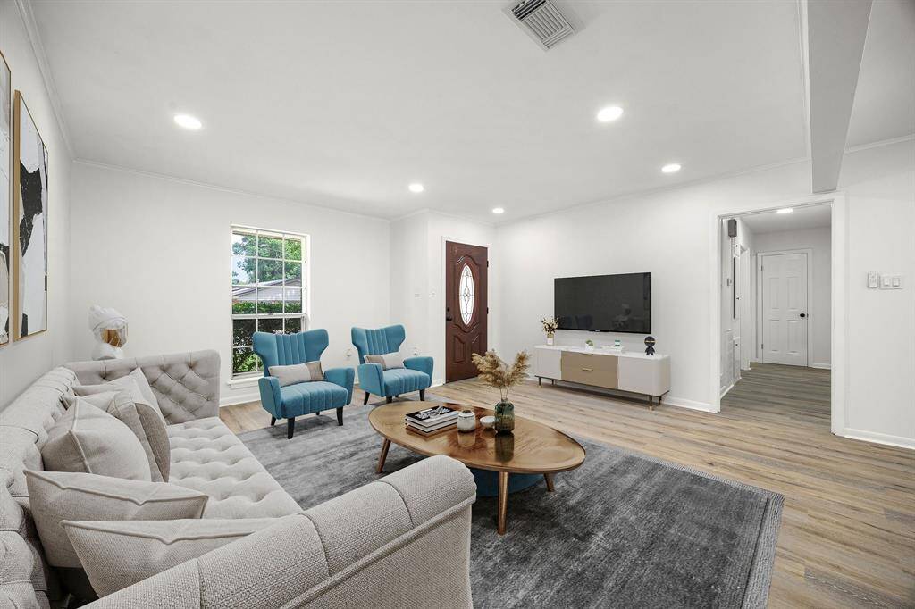 Spacious living room with modern decor features a cozy sectional sofa, two blue armchairs, and a sleek TV stand. The room is bright with natural light from the window and has recessed lighting. Light wood floors and neutral walls create an inviting atmosphere.