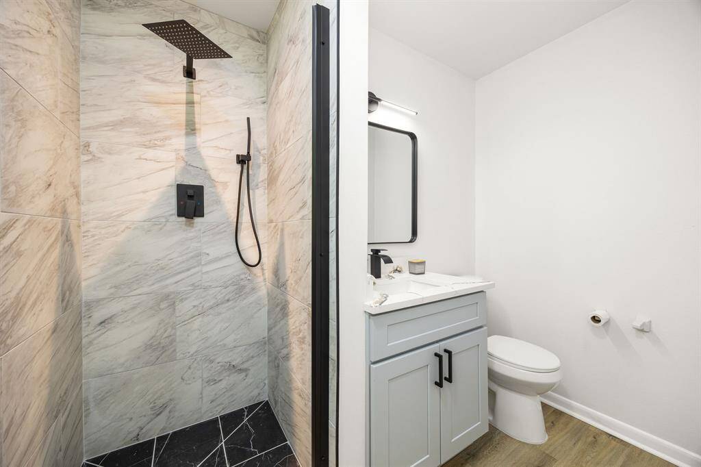 This bathroom features a sleek modern design with a marble-tiled shower, a rainfall showerhead, and a handheld attachment. There's a stylish vanity with a large mirror and ample under-sink storage, alongside a standard toilet. The space is well-lit and has a clean, minimalist aesthetic.