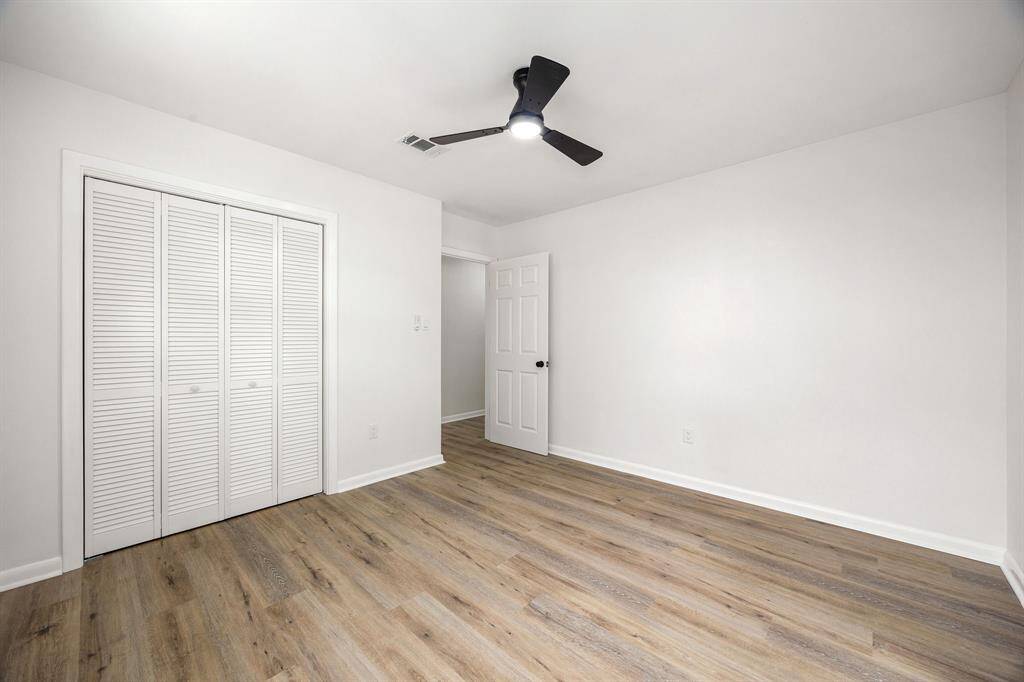 This room features clean white walls and wood-look flooring, creating a modern and inviting space. It includes a ceiling fan for comfort and a closet with louvered doors for storage.