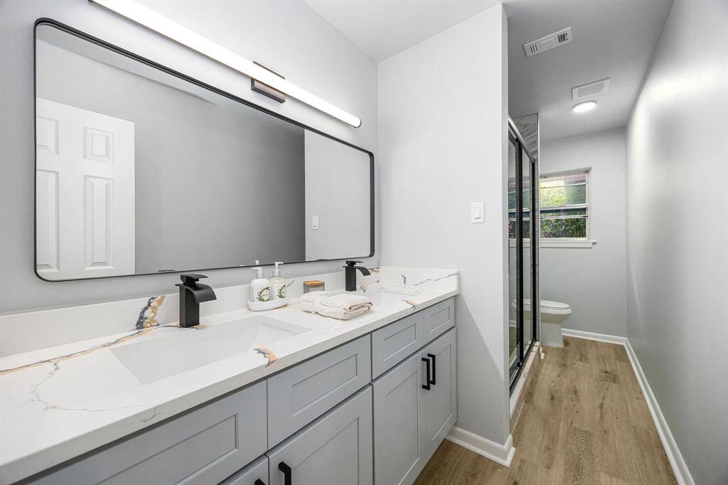 This modern bathroom features a sleek design with a large mirror, dual sinks, and stylish faucets. It includes ample cabinet space, a glass-enclosed shower, and wood-look flooring for a clean, contemporary feel.