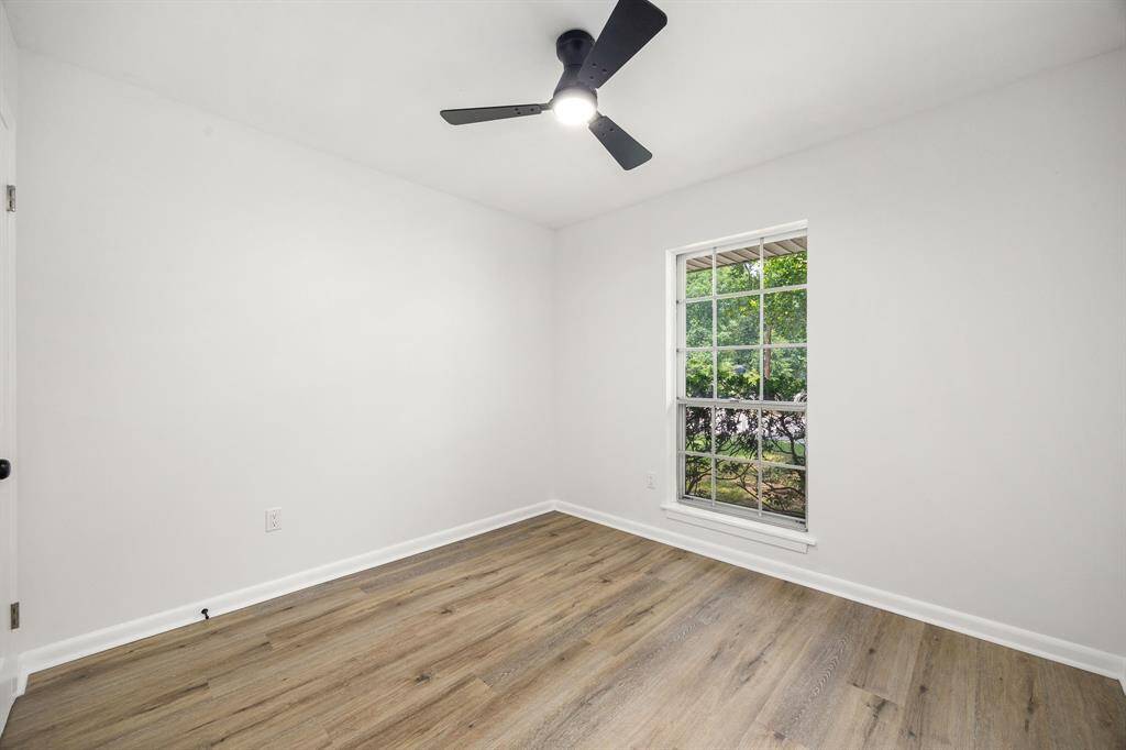 This room features fresh white walls, light wood flooring, and a large window that offers natural light and views of greenery. It includes a modern ceiling fan, creating a bright and airy space.