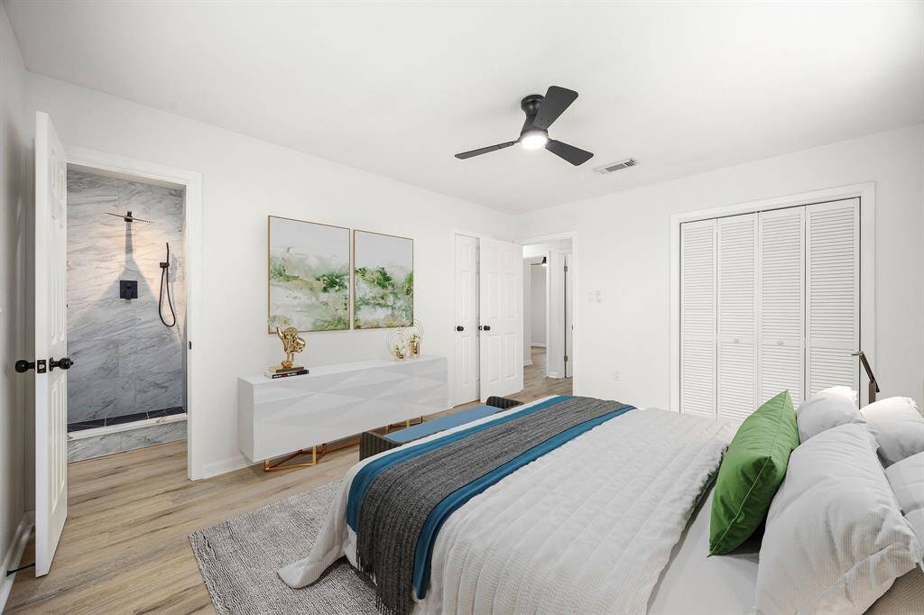 This bedroom features a modern design with light wood flooring and a neutral color palette. It includes a ceiling fan, a large closet with louvered doors, and an en-suite bathroom showcasing a sleek, tiled shower. The space is stylishly furnished with simple decor and bedding.
